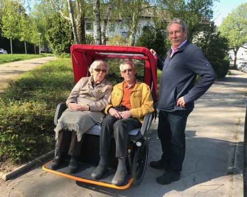 Eine Seniorin und ein Senior sitzen in einem Lastenrad, daneben steht in Mann und lächelt